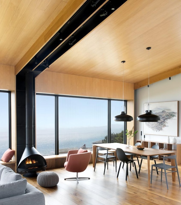 Ein modernes Interieur mit einem großen Kamin, Essbereich und Panoramafenstern mit Blick auf eine Landschaft.