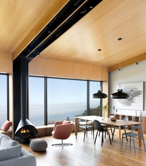A modern interior with a large fireplace, dining area, and panoramic windows overlooking a landscape.