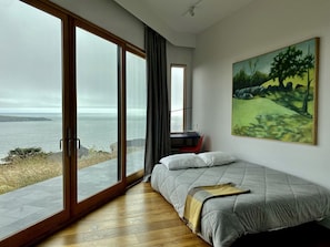 Ein Schlafzimmer mit einem großen Schiebeglasfenster, das Meerblick bietet, einem Bett mit Decke und einem Gemälde an der Wand.