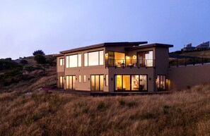 A modern two-story building with large windows and a balcony, situated on a grassy hill.