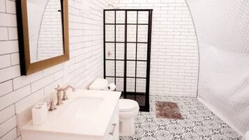 A bathroom with a white tiled wall, a large mirror, a sink with a faucet, a toilet, and a shower enclosure.