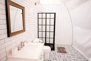 A bathroom with a white tiled wall, a large mirror, a sink with a faucet, a toilet, and a shower enclosure.
