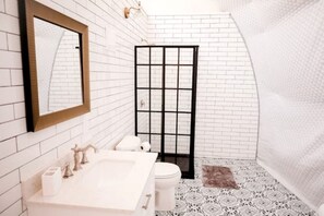 Ein Badezimmer mit weiß gefliester Wand, einem großen Spiegel, einem Waschbecken mit Wasserhahn, einer Toilette und einer Duschkabine.