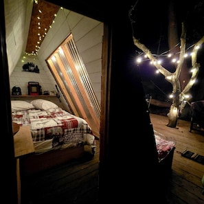 A cozy bedroom with a slanted ceiling, a bed with a checkered blanket, a small table with a radio, and string lights.