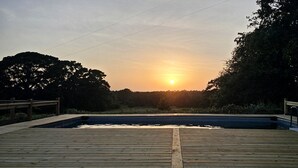 Une terrasse en bois avec une piscine, entourée d’arbres, et un coucher de soleil en arrière-plan.
