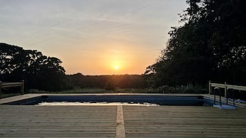 Eine Holzterrasse mit Schwimmbad, umgeben von Bäumen und einem Sonnenuntergang im Hintergrund.