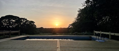 A wooden deck with a swimming pool, surrounded by trees and a sunset in the background.