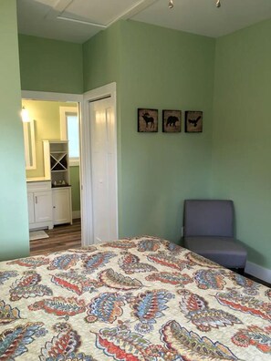 A bedroom with a patterned bedspread, a gray chair, and a door leading to a bathroom.