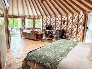A cozy interior of a yurt with a bed, a sofa, a television, and a wooden table. - Incredible Spacious Yurt with Hot Tub and Fire Pit Surrounded by Nature for an Unforgettable Glamping Experience in Woodstock, Maine (Woodstock)