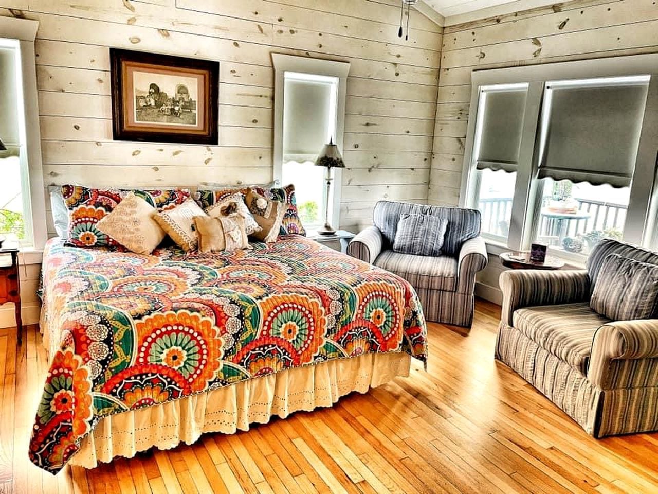 A cozy room with a wooden floor, a large bed with a colorful quilt, a striped armchair, and a framed picture on the wall.