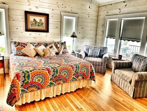 A cozy room with a wooden floor, a large bed with a colorful quilt, a striped armchair, and a framed picture on the wall.