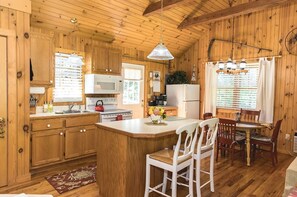 A wooden cabin interior with a kitchen, dining area, and a large window.