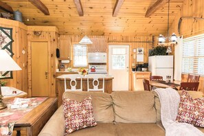 Un interior de cabaña acogedor con techo de madera, un sofá con cojines estampados, una mesa de comedor y una zona de cocina con estufa y refrigerador.