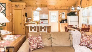 A cozy cabin interior with a wooden ceiling, a sofa with patterned cushions, a dining table, and a kitchen area with a stove and refrigerator.