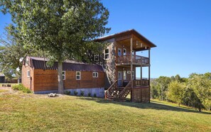 Une cabane en bois à deux étages, dotée d’un balcon, d’un escalier et d’un grand arbre en arrière-plan.