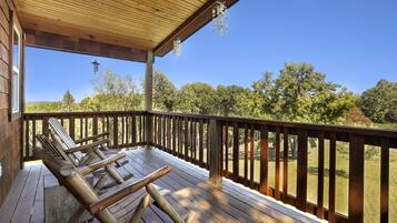 Una terraza de madera con barandilla, zona cubierta y vista a árboles y colinas.