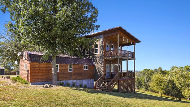 A two-story wooden cabin with a balcony, a staircase, and a large tree in the background.