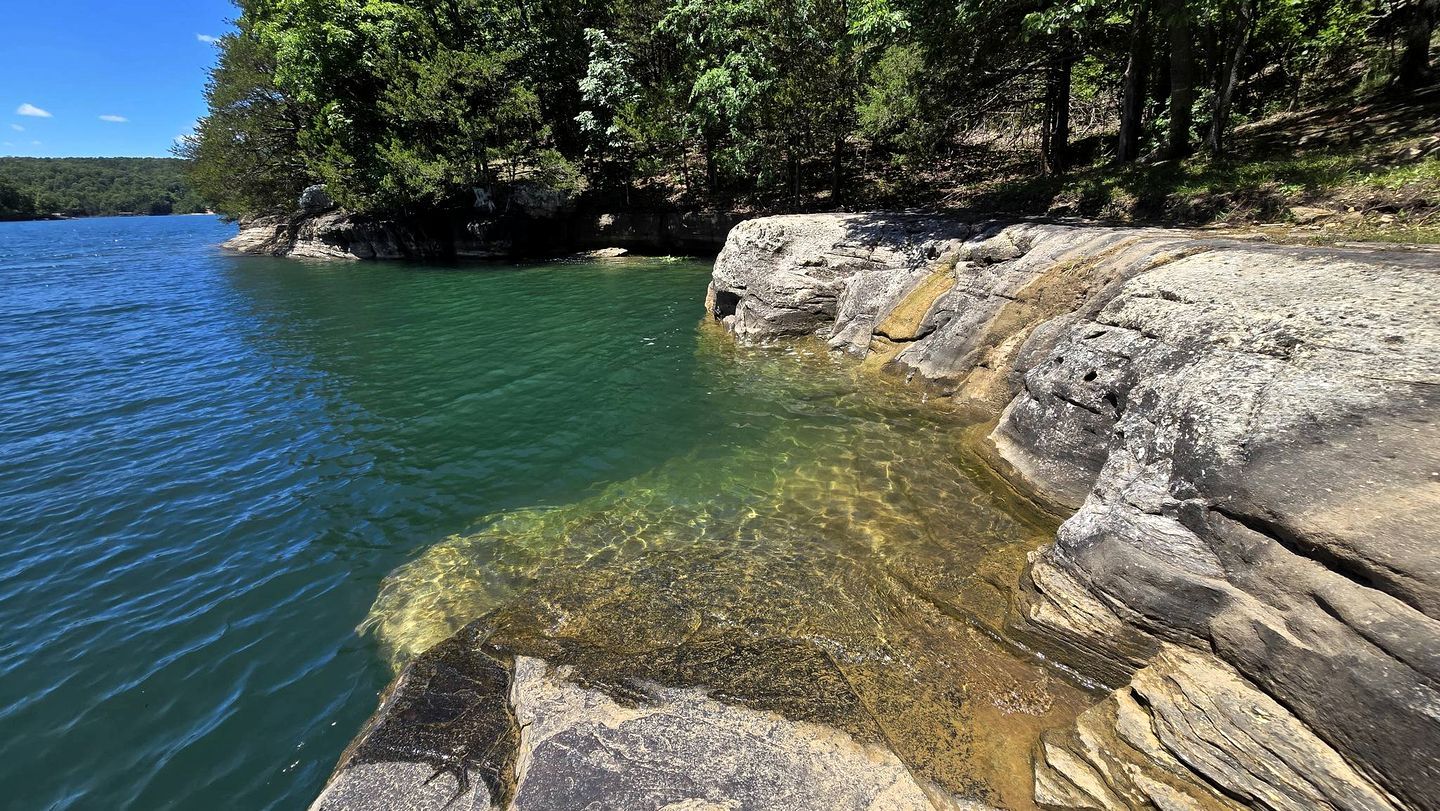 A natural landscape with a clear water body, rocky formations, and dense greenery.
