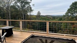 Una terrassa de fusta amb taula i cadires, envoltada per una tanca de filferro, amb vista a un paisatge forestal.