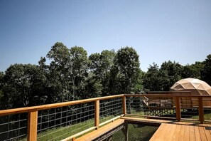 Een houten terras met een metalen hekwerk, een overdekte zithoek en uitzicht op dichte bomen.