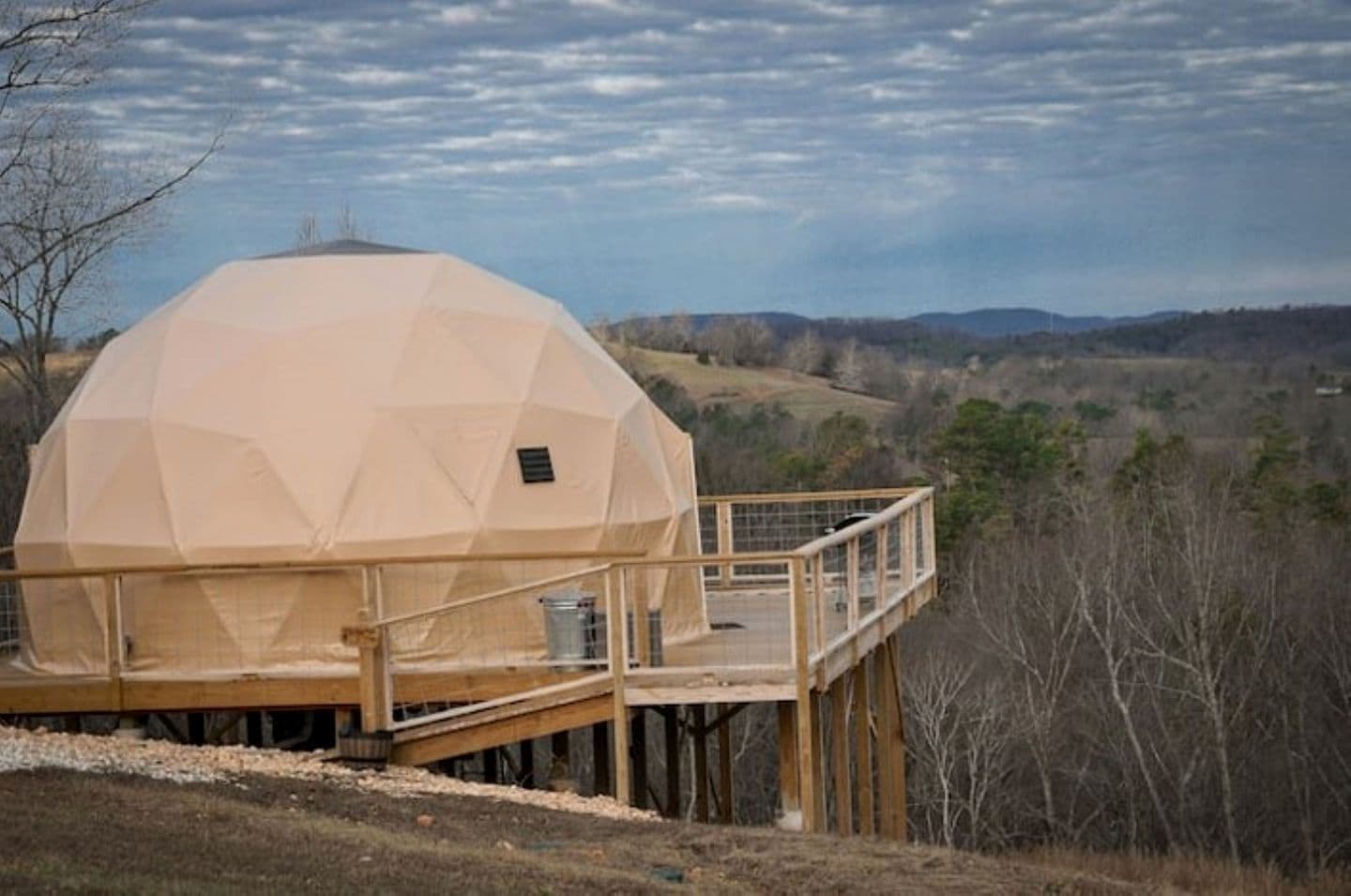 Une structure en forme de dôme géodésique, dotée d’une terrasse et d’une rambarde en bois, qui se détache sur un paysage rural vallonné et parsemé d’arbres.