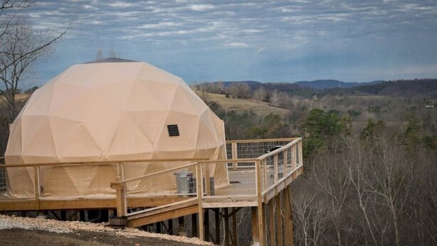 Una estructura de domo geodésico con terraza y barandilla de madera, en un paisaje rural con colinas y árboles.