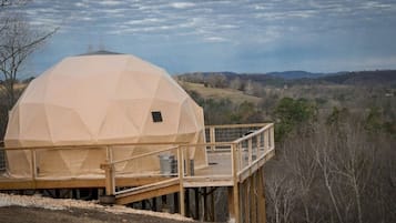 Une structure en forme de dôme géodésique, dotée d’une terrasse et d’une rambarde en bois, qui se détache sur un paysage rural vallonné et parsemé d’arbres.