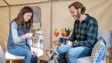 Dos personas sentadas en una tienda de campaña con un perro, una mesa con bebidas y un cojín.