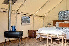 A tented room with a bed, a small table, a heater, and a window with a forest view.