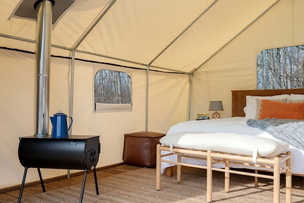 A tented room with a bed, a small table, a heater, and a window with a forest view.