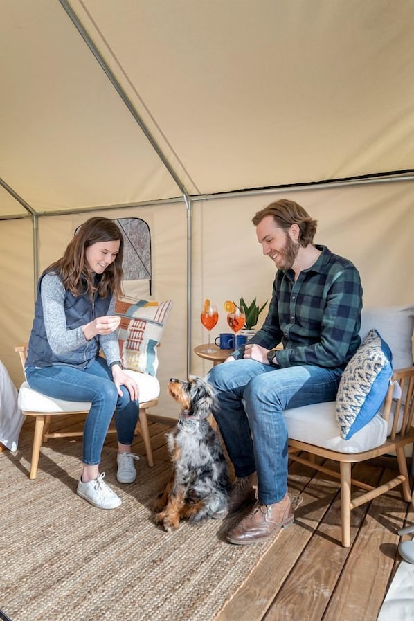 Zwei Personen sitzen in einem Zelt, mit einem Hund, einem Tisch mit Getränken und einem Kissen.