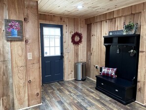 A wooden interior with a door, a trash can, a bench with a 'Merry' pillow, and a wreath on the wall.