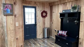A wooden interior with a door, a trash can, a bench with a 'Merry' pillow, and a wreath on the wall.