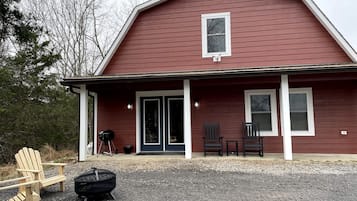 A red building with a porch, two chairs, a grill, and a fire pit.