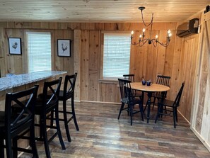 A wooden-paneled room with a kitchen area, dining table, chairs, and framed pictures on the walls.