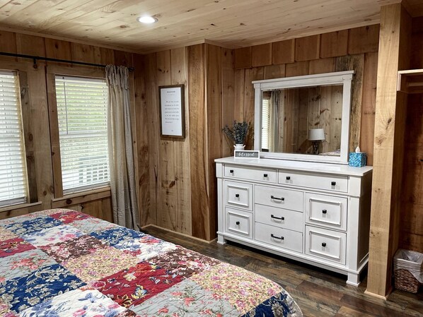 A bedroom with wooden walls, a white dresser, a large mirror, and a floral bedspread.