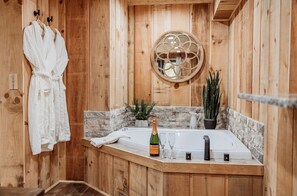 A wooden cabin bathroom with a bathtub, a robe hanging on a rack, a bottle of champagne, and a potted plant.