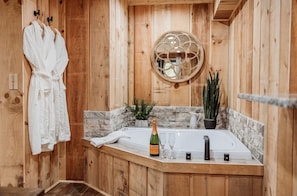 A wooden cabin bathroom with a bathtub, a robe hanging on a rack, a bottle of champagne, and a potted plant.