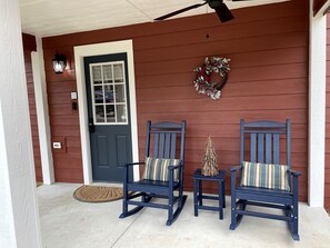 A porch with two rocking chairs and a small table, a door, and a wreath on the wall.