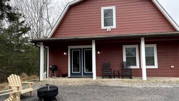 A red building with a porch, two chairs, a grill, and a fire pit.
