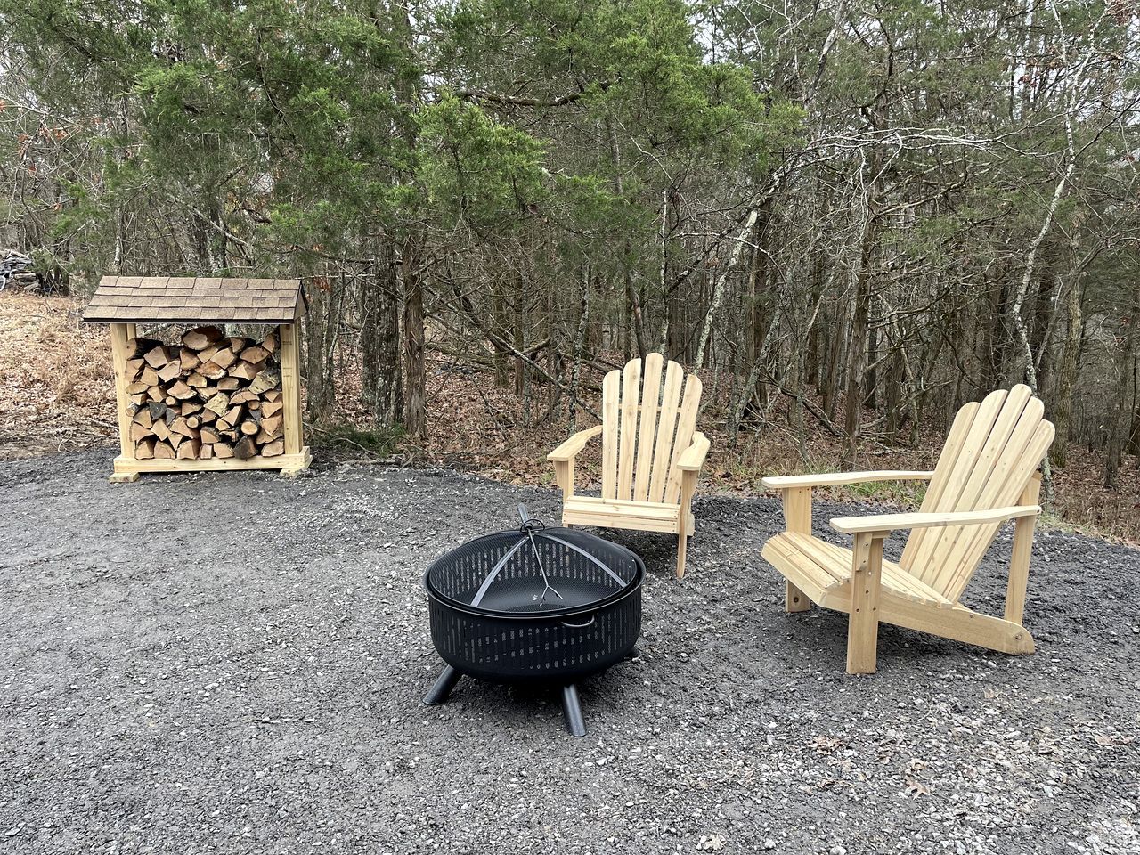 A forest setting with a fire pit, two wooden Adirondack chairs, and a stack of firewood.