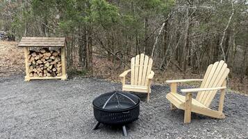 A forest setting with a fire pit, two wooden Adirondack chairs, and a stack of firewood.