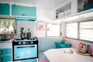 A compact kitchen with teal cabinets, a vintage stove, a window with blinds, and a pink bench. - Vintage Camper Rental in Franklin for Couples Getaway in the Mountains in North Carolina (Franklin)