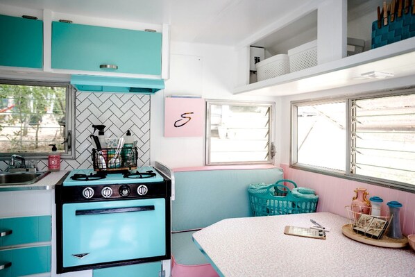 Una cocina compacta con gabinetes color turquesa, una estufa antigua, una ventana con persianas y un banco rosa.