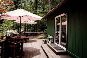 A wooden deck with outdoor furniture and umbrellas, a green building with glass doors, and a forested background. - Vintage Camper Rental in Franklin for Couples Getaway in the Mountains in North Carolina (Franklin)