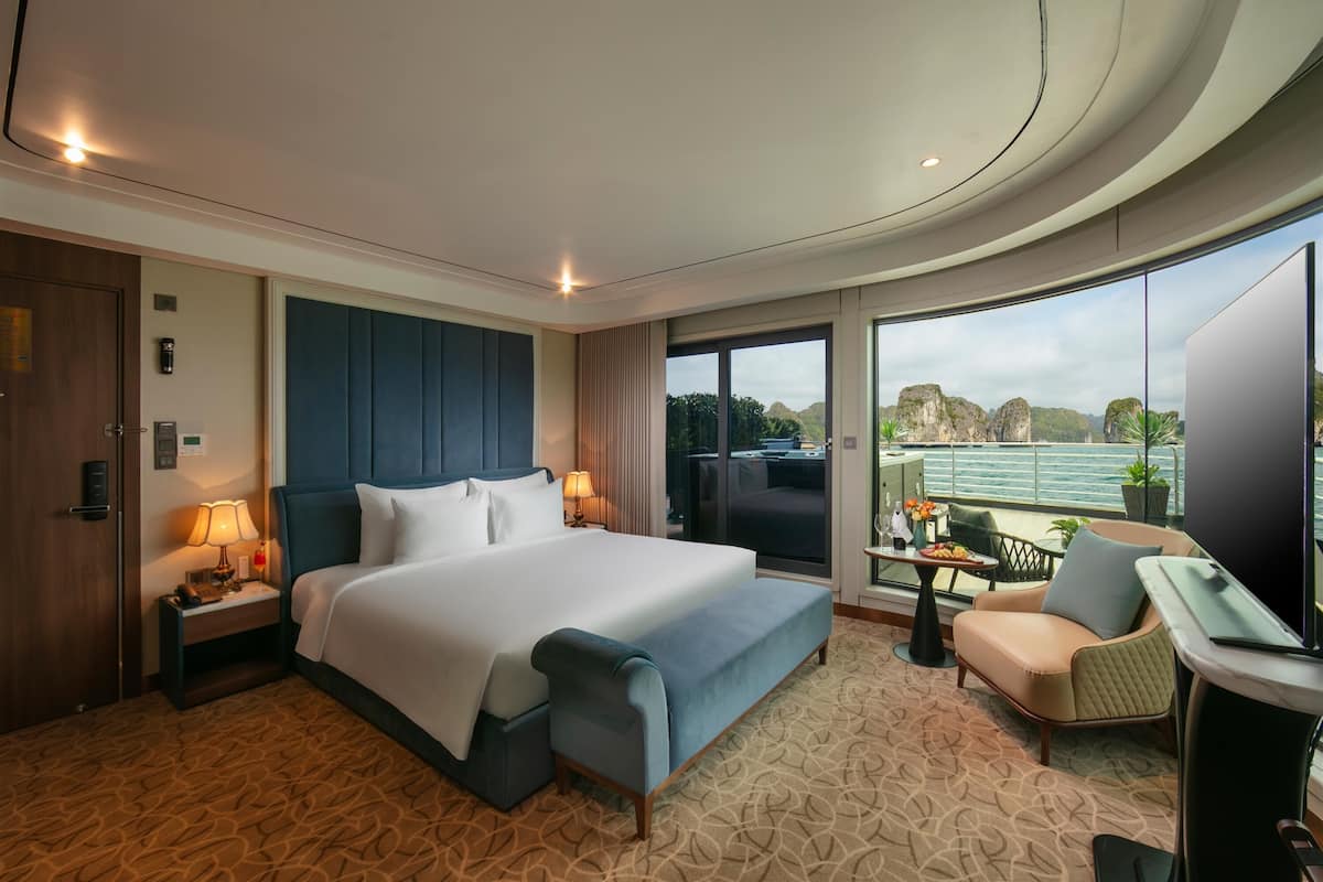 Ein modernes Hotelzimmer mit einem großen Bett, einer Sitzecke mit Sofa und Sesseln, einem kleinen Tisch und einem großen Fenster mit Blick auf das Meer und die Berge.