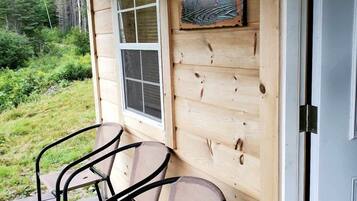 A wooden cabin with a porch, two metal chairs, a window, and a welcome sign.