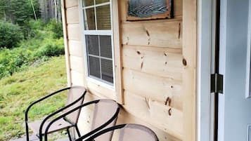 A wooden cabin with a porch, two metal chairs, a window, and a welcome sign.