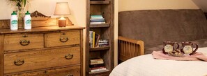 A bedroom with a wooden dresser, a bedside table with a lamp, a bookshelf, a bed with a checkered blanket, and two mugs on the bed. - Lovely Suite Rental for Two at Countryside Bed and Breakfast in Warren, Vermont (Warren)
