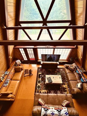 A spacious loft-style living room with high ceilings, a large TV, and a view of a forest. - Spacious Well Furnished A-Frame with Sauna in Union Dale, Pennsylvania (Union Dale)